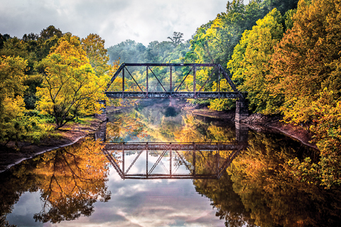 "Train's A'Comin'" by Debra and Dave Vanderlaan... Deep in the heart of the Appalachian Mountains, this vintage trestle spans the Valley River in the little country town of Murphy, North Carolina. Captured in the early morning just after sunrise, when the fog and mist was hanging over the still water of this pretty stream, the reflections are perfect. The trees are lush with early fall foliage surrounding the old metal railroad trestle. The granite block pilon supports are on the edge of the river which runs much higher in the Spring and Summer. In the autumn months, the dams are opened and the water recedes to expose the clay banks of the riversides. Clouds hang low, joining the morning fog and touching the autumn color in the oak, maple, willow, sycamore, and poplar trees in the Blue Ridge mountain forest.